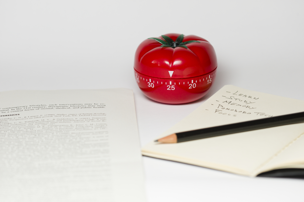 Ein Kurzzeitmesser in Tomatenform für die Pomodoro-Technik, ein geöffnees Notizbuch mit Stift und ein ausgdruckter Text.